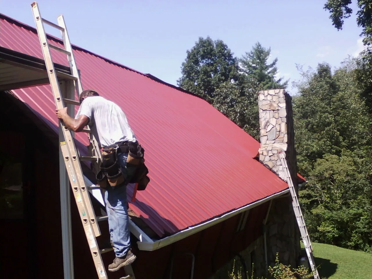 Roofer on a ladder beside a red metal roof and stone chimney — Soffit & Fascia Repair by Roofing Brothers in Wabash